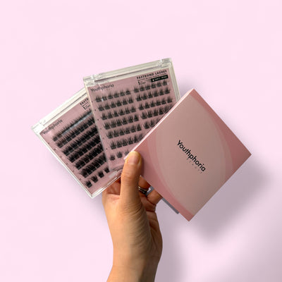 Hand holding a set of false eyelashes with 'Youthphoria' packaging on a light pink background
