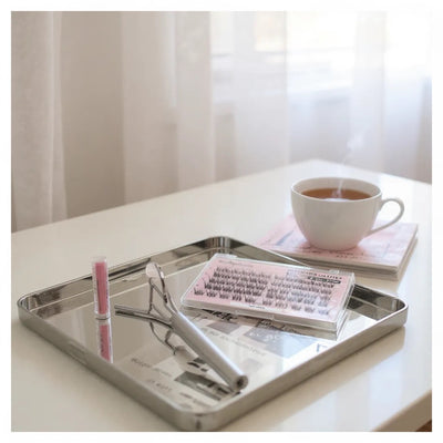 Metal tray with a cup of tea, sunglasses, false lashes and a book on a table.