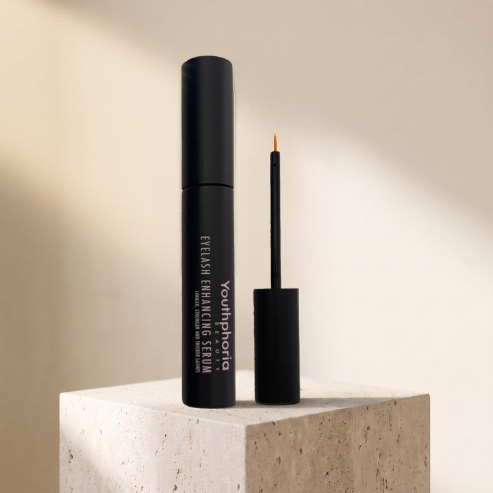 Youthphoria Eyelash Growth and Repair Serum bottle and applicator brush standing on a travertine block in window light