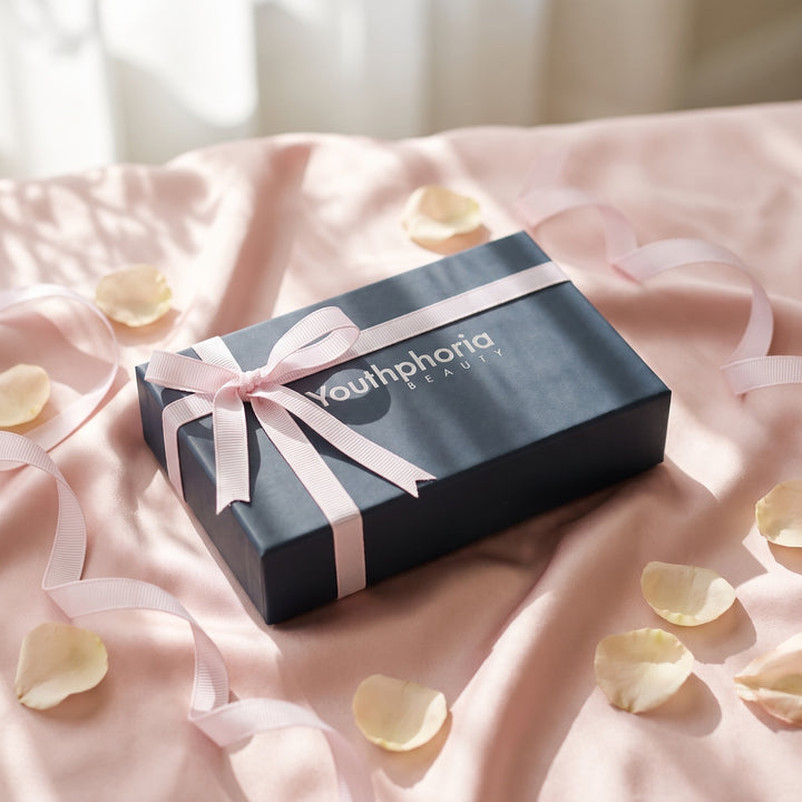Youthphoria Beauty navy gift box tied with a pale pink grosgrain ribbon bow, resting on blush pink silk in soft morning light with scattered petals
